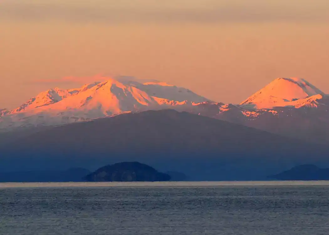 Distant snow-capped mountains glow with pink sunrise light, overlooking a calm blue lake—a serene scene that beautifully captures the tranquility of a Taupō lifestyle estate investment. A small, dark island sits in the foreground, while the sky is softly tinted with warm hues.