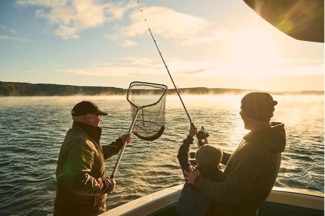 Three people on a boat are fishing at sunrise, discovering firsthand why Taupō is renowned for its natural beauty. One holds a fishing net, while another helps a child reel in a fishing rod over the water. Mist rises above the lake under a bright sun.