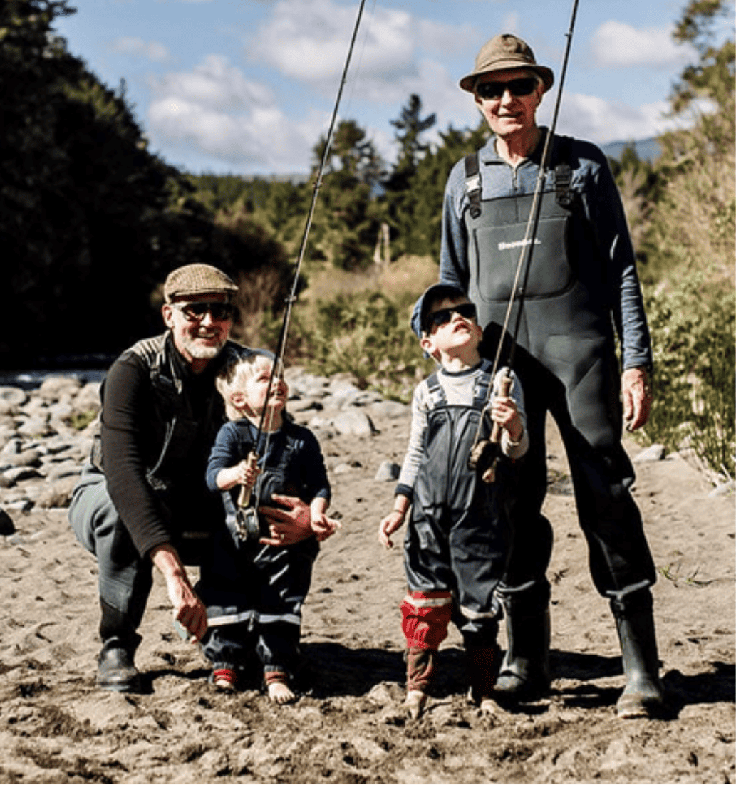 Two adults and two children, all in waders and hats, stand with fishing rods on a rocky riverbank surrounded by trees, enjoying a sunny day outdoors together—capturing the relaxed Taupō lifestyle.
