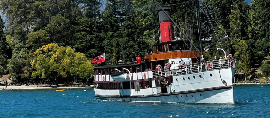 A white steamship with a red smokestack sails on a blue lake, surrounded by tall trees and mountains under a clear sky—a serene scene that embodies why Taupō is celebrated for its breathtaking beauty.