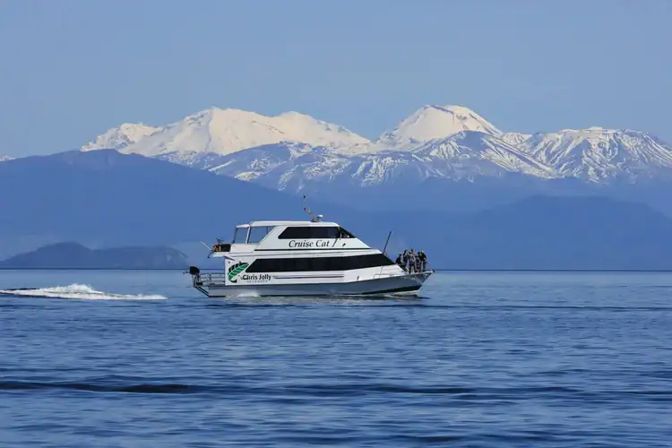 A white cruise boat travels across calm blue water with snow-capped mountains and a clear sky in the background, showcasing why Taupō is renowned for its breathtaking natural beauty.