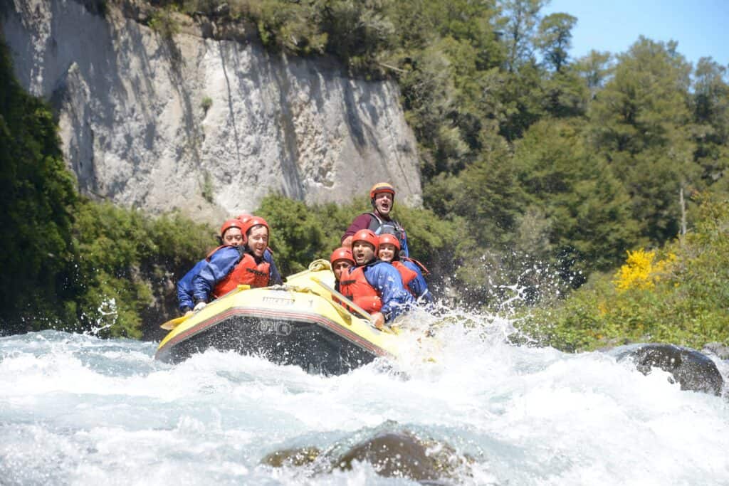 Six people wearing helmets and life jackets are whitewater rafting on a river surrounded by trees and cliffs, with water splashing around them—a thrilling adventure that truly captures the essence of the Taupō lifestyle.