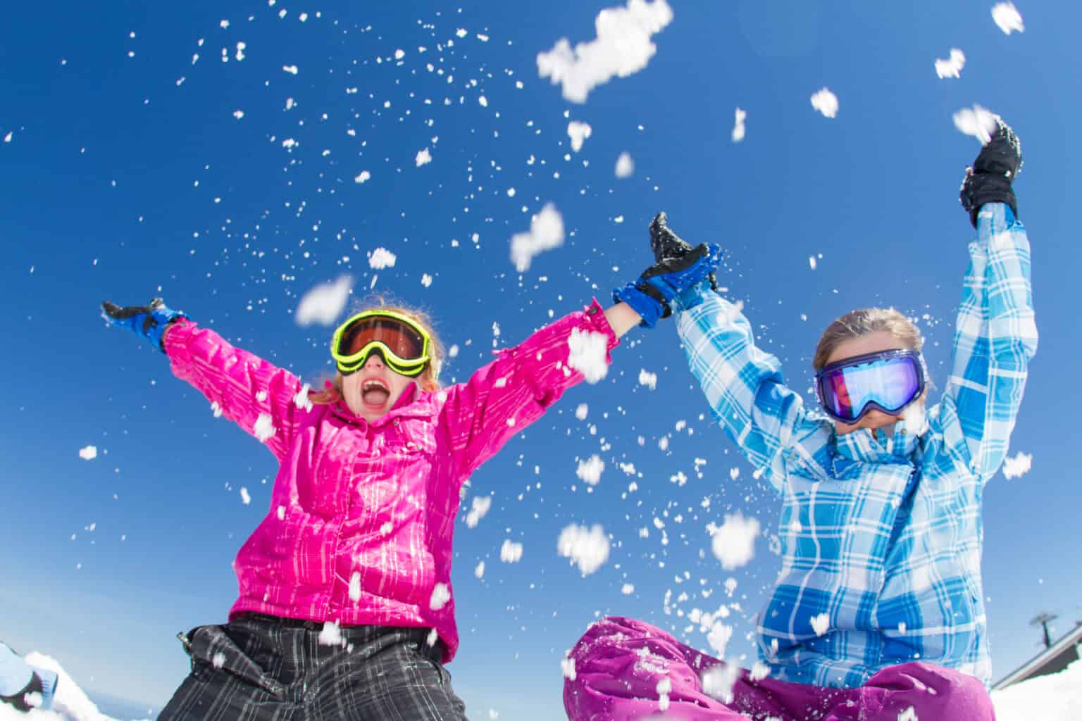 Two children in colorful winter jackets and goggles joyfully throw snow into the air, smiling under a bright blue sky.