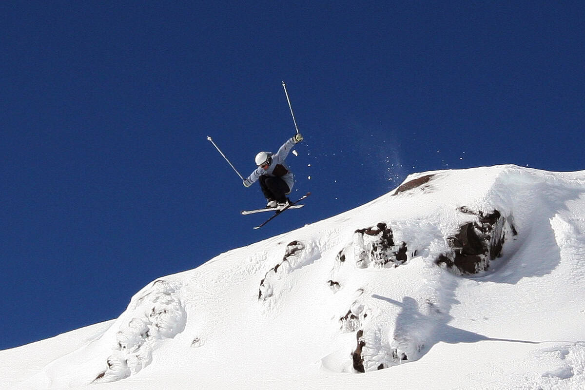 A skier wearing a helmet is mid-air above a snowy slope, holding ski poles, with clear blue sky in the background.