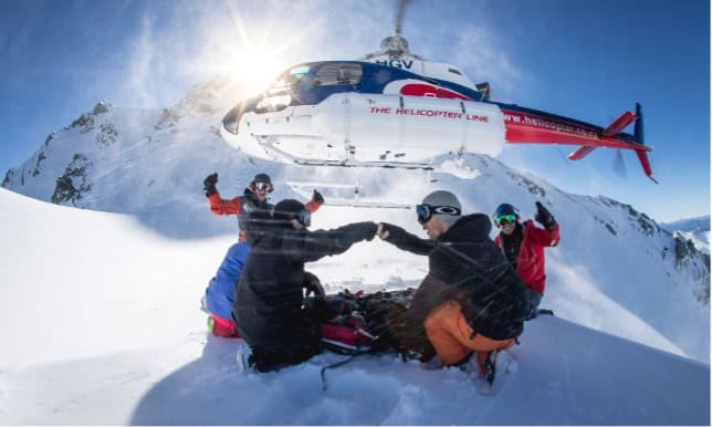 Four people in snow gear fist bump while sitting on snowy ground as a helicopter hovers close by in a sunny, mountainous area. Snow swirls in the air, and another mountain peak is visible in the background.
