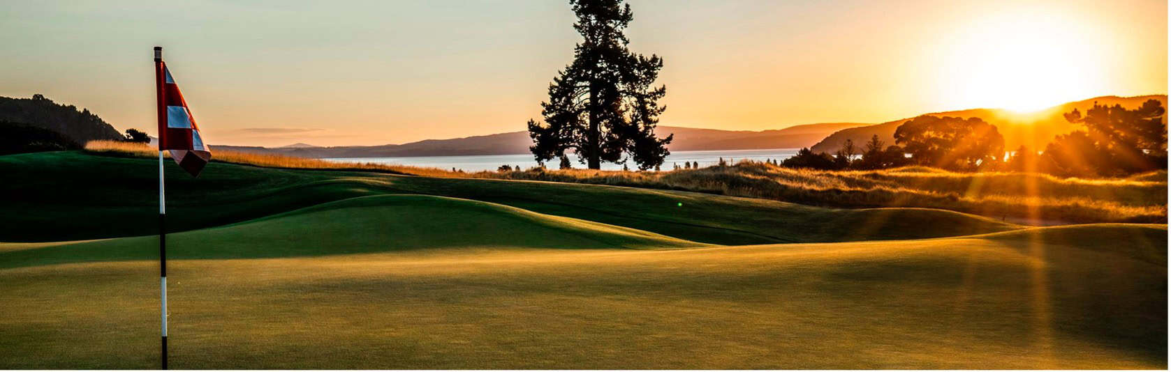 A golf course at sunset with a flagstick in the foreground, rolling green hills, silhouetted trees, and sunlight shining over the landscape near a body of water—an idyllic scene that captures the essence of a Taupō lifestyle estate investment.