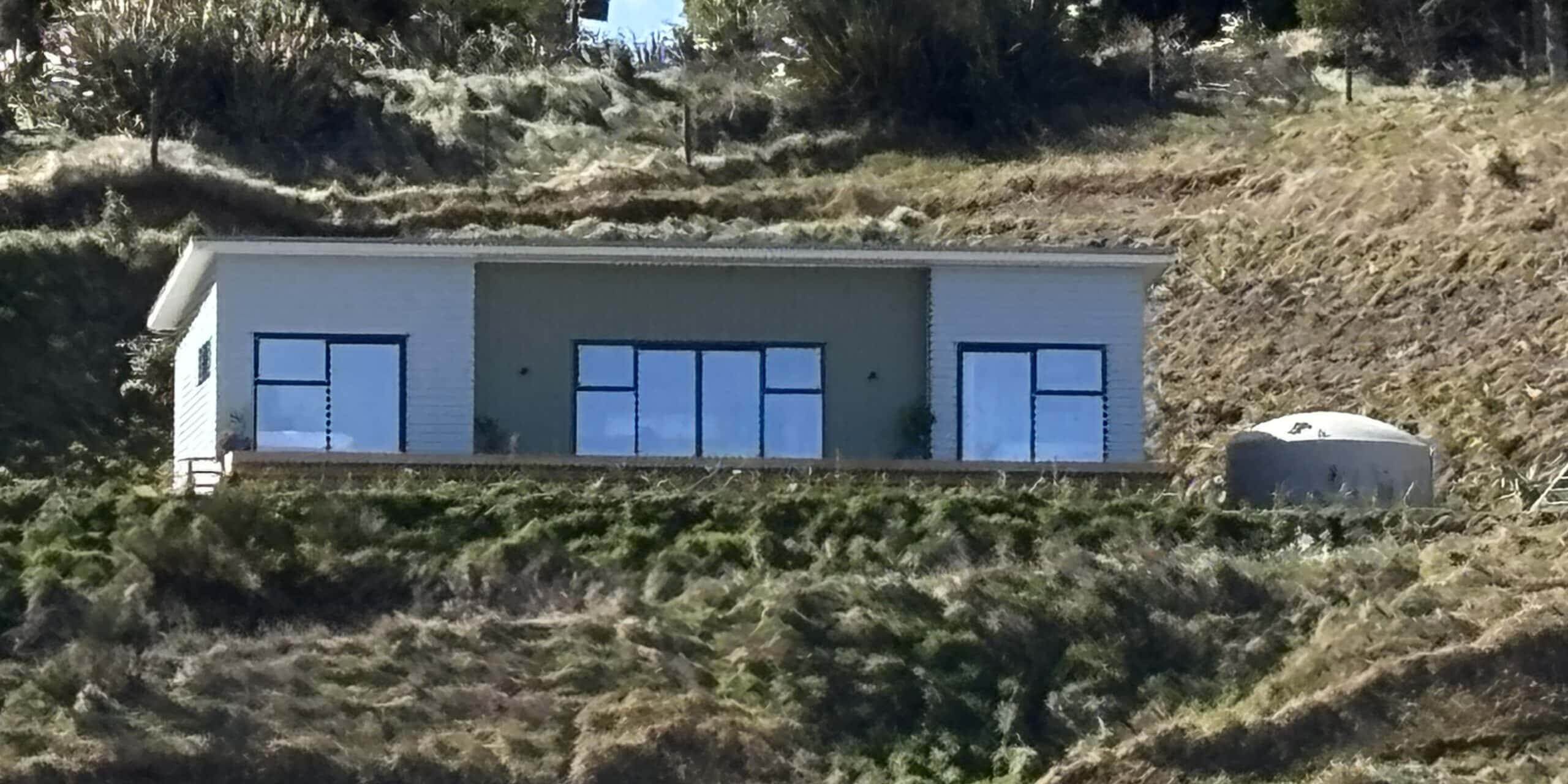A modern, single-story house with large windows sits on a grassy hillside. A small water tank is located to the right of the house, surrounded by sparse vegetation and trees.