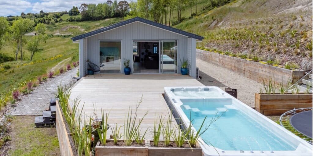A modern house with large sliding glass doors sits in a green hilly landscape, forming the centerpiece of this Taupō lifestyle estate for sale. A long wooden deck features a swim spa pool, potted plants, and a trampoline, with garden beds and stone paths surrounding the area.